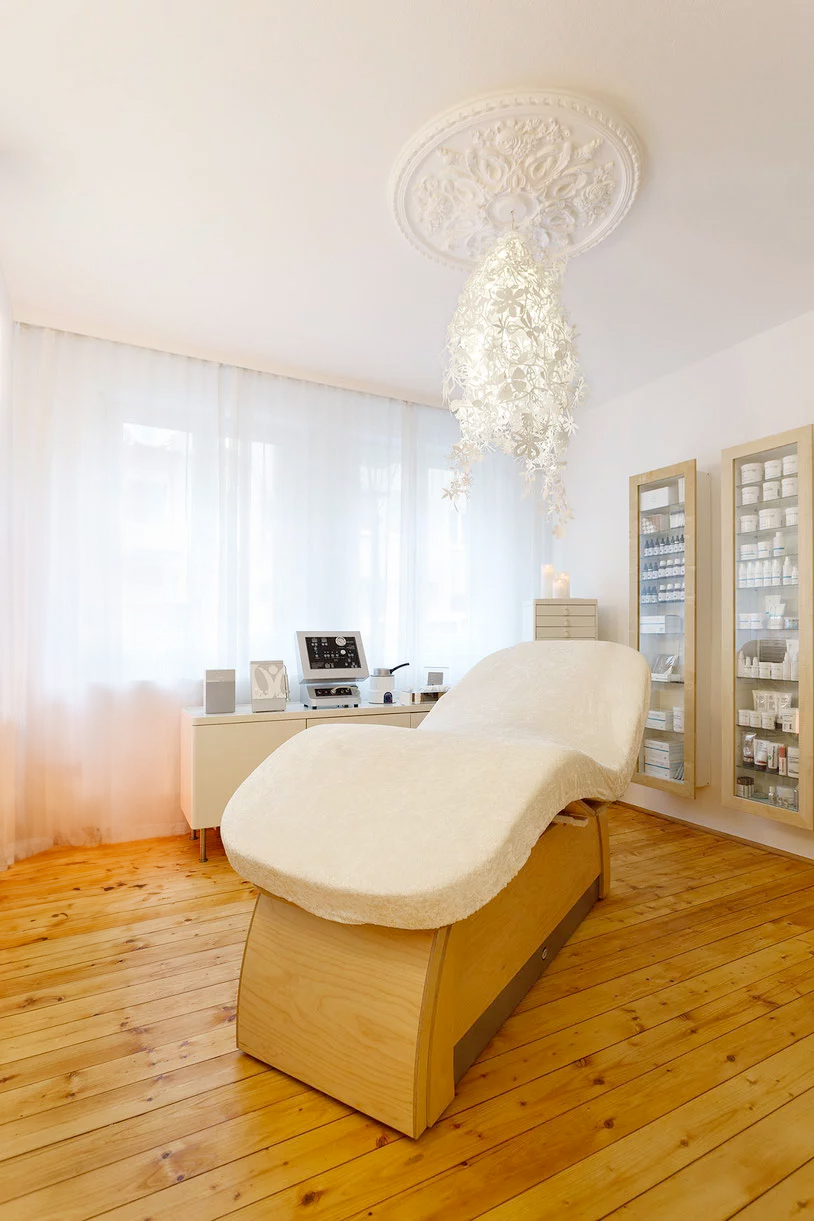 Aufnahme vom Behandlungsraum, eine große bequeme Liege mit cremeweißem Bezug steht diagonal in einem hellen Raum mit Holzdielen, weißen Wänden und weißen Vorhängen. Im Hintergrund zwei Wandvitrinen und ein Sideboard mit kosmetischen Arbeitsgeräten. Über der Liege an der Decke eine große Stuckrosette mit einer weißen floralen Deckenlampe